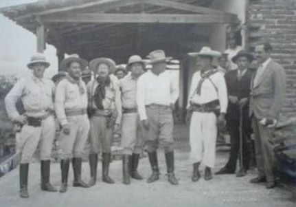 Grupo de hombres con sombrero, uniforme y cananas, de pie frente a una construcción con tejado, junto a dos hombres de traje.